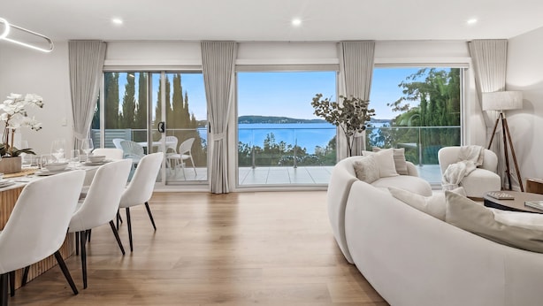 A modern living room with a large window, a dining area with white chairs, and a sofa with a view of the outdoors. - Bayview Luxe Retreat | Luxury Central Coastal Escape with Panoramic Water Views (Tascott)