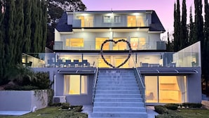 A modern two-story house with a staircase leading to the entrance, surrounded by well-maintained lawns and tall trees. - Bayview Luxe Retreat | Luxury Central Coastal Escape with Panoramic Water Views (Tascott)
