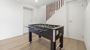 A modern room with a foosball table, wooden flooring, white walls, and a staircase. - Bayview Luxe Retreat | Luxury Central Coastal Escape with Panoramic Water Views (Tascott)