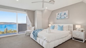 A bedroom with a large bed, a ceiling fan, a nightstand, a chair, and a view of the sea. - Bayview Luxe Retreat | Luxury Central Coastal Escape with Panoramic Water Views (Tascott)