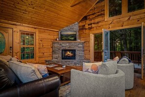 A log cabin interior with a stone fireplace, wooden walls, and a view of the forest through a glass door. - Luxury Cabin for Riders Hikers & Anglers +Hot Tub (Pisgah Forest)