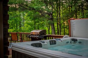 Ein Whirlpool und ein Grill auf einer Holzterrasse mit Wald im Hintergrund.