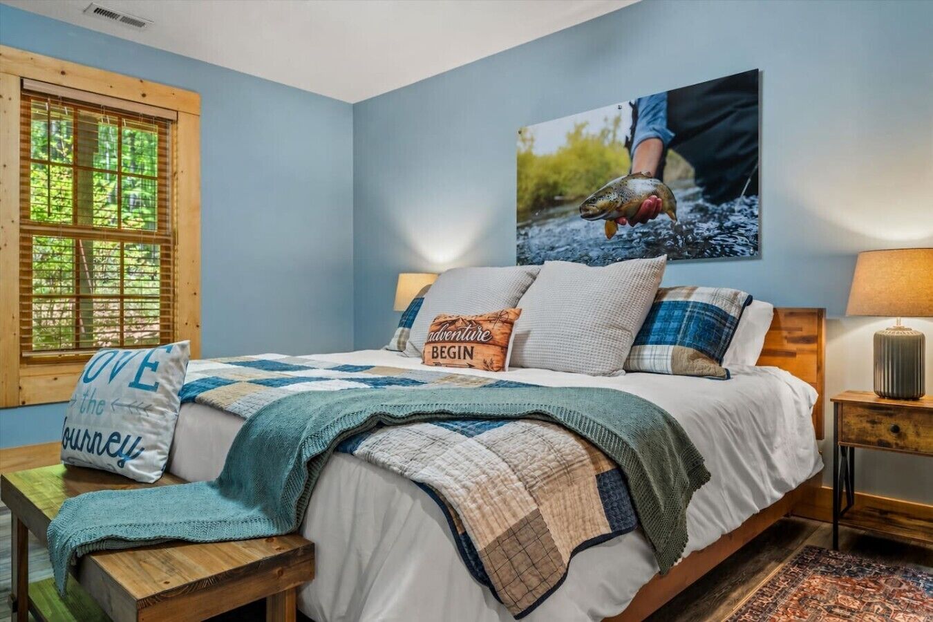Un dormitorio con cama, almohadas, manta, ventana, un cuadro y una mesita de noche.