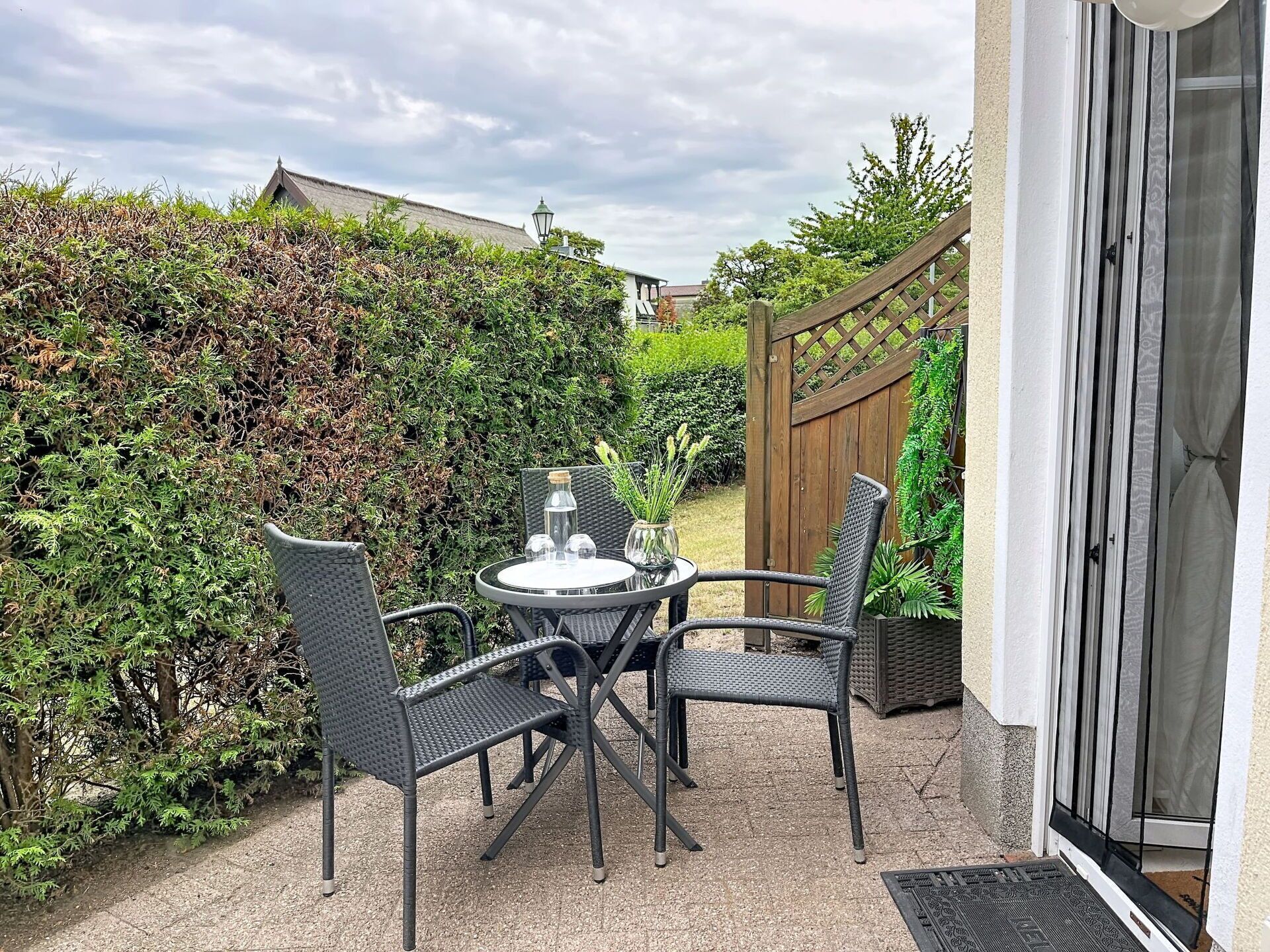 Eine Terrasse mit rundem Tisch und vier Stühlen, umgeben von dichter Bepflanzung und einem Holzzaun.