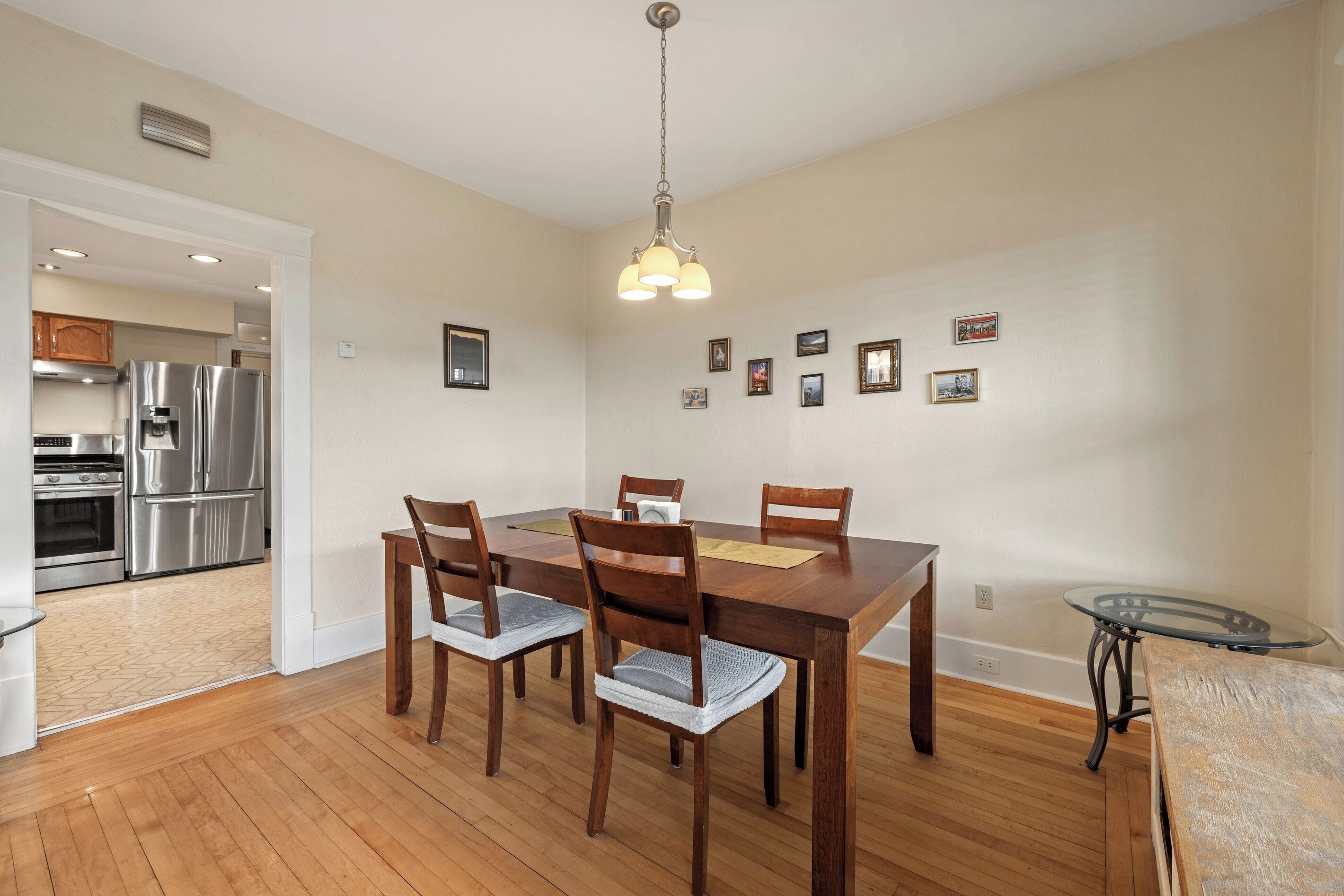 A dining area with a wooden table and chairs, a hanging light fixture, and a kitchen in the background.