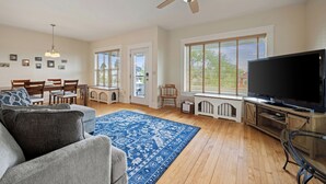 A living room with a ceiling fan, a large TV, a blue patterned rug, and wooden flooring. - U-District 1 Bedroom Craftsman w/ Fenced Yard (Butte)