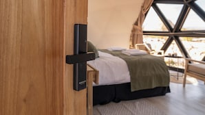 Eine Holztür mit einem schwarzen Gegenstand, ein Bett mit einer grünen Decke und Blick durch ein Fenster.