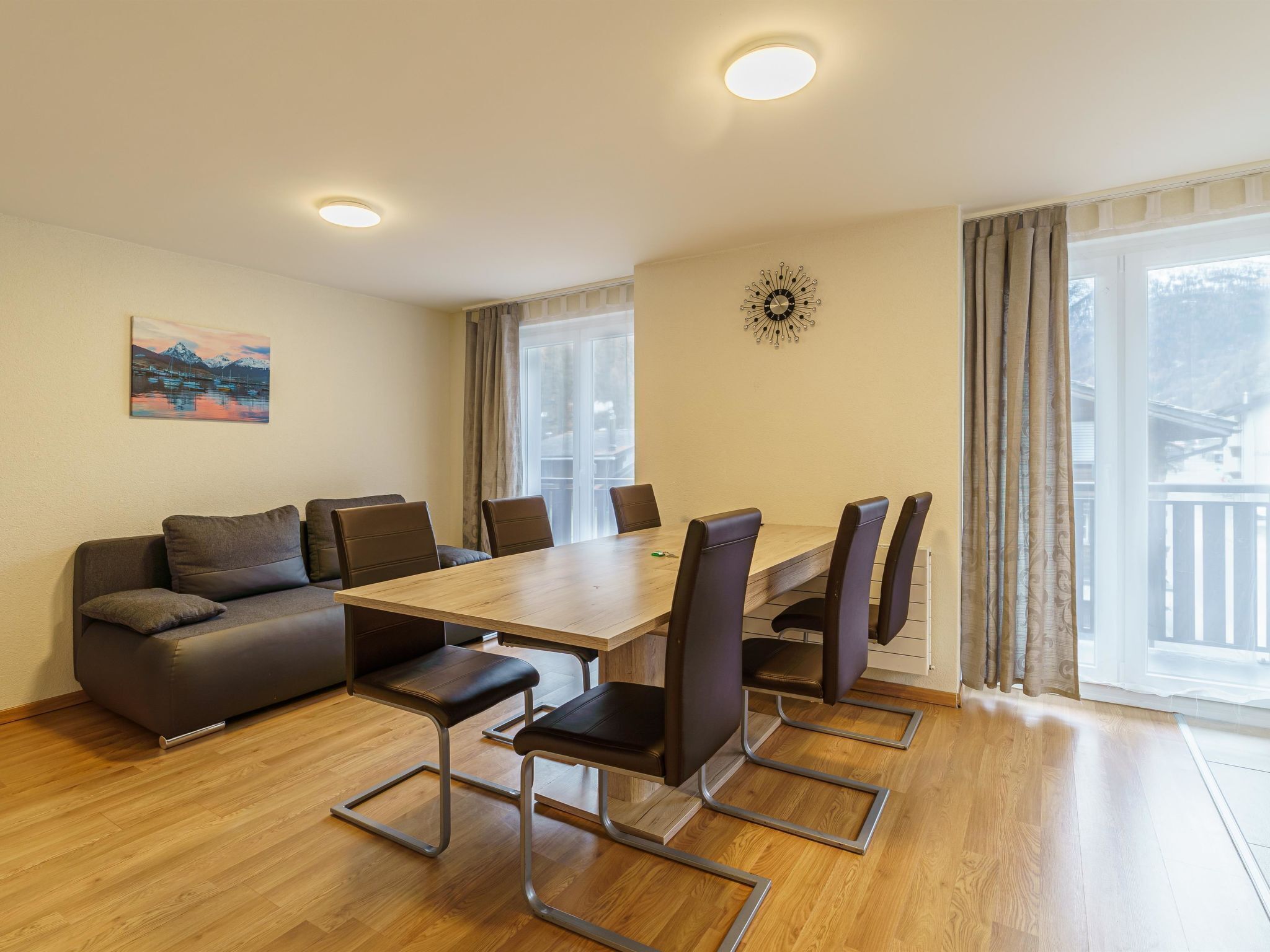 Ein modernes Wohnzimmer mit einer Couch, einem Esstisch und Stühlen. An der Wand hängt ein Gemälde und eine Uhr ist zu sehen.