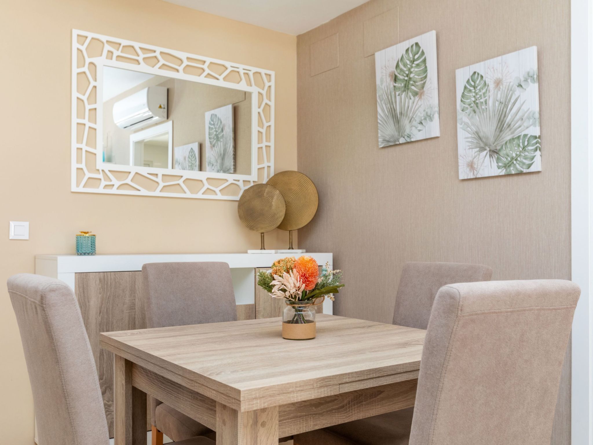 Un comedor con una mesa de madera, sillas color beige, un jarrón con flores, un espejo decorativo e imprime de plantas colgadas en la pared.