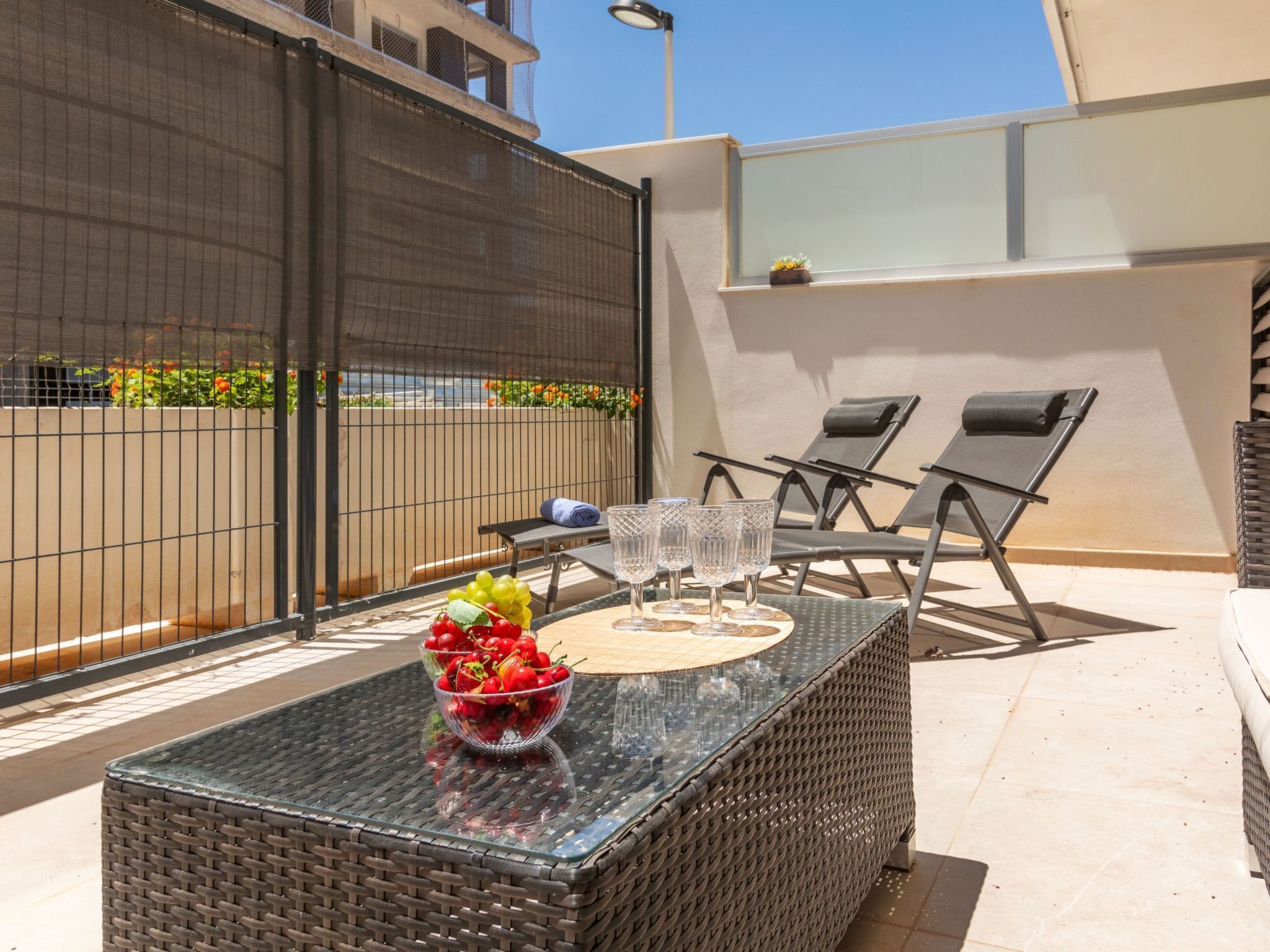 A balcony with a wicker table, two lounge chairs, and a glass-top table with a bowl of fruit and glasses.