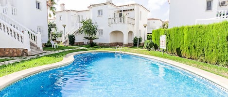 A swimming pool with a curved edge, surrounded by white buildings with arched windows and a balcony.