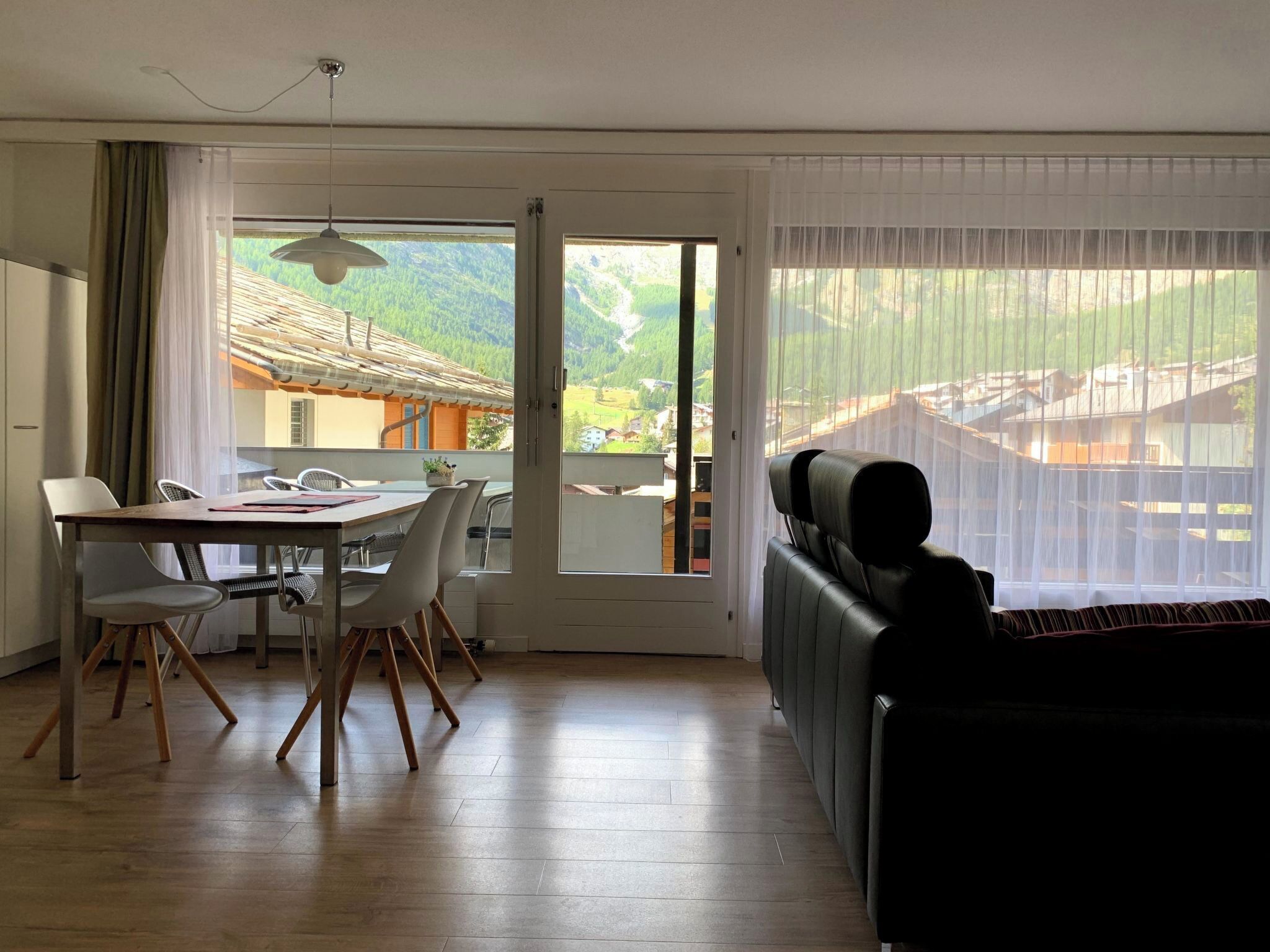 A room with a dining table, chairs, a sofa, and large windows with a view of a mountainous landscape.