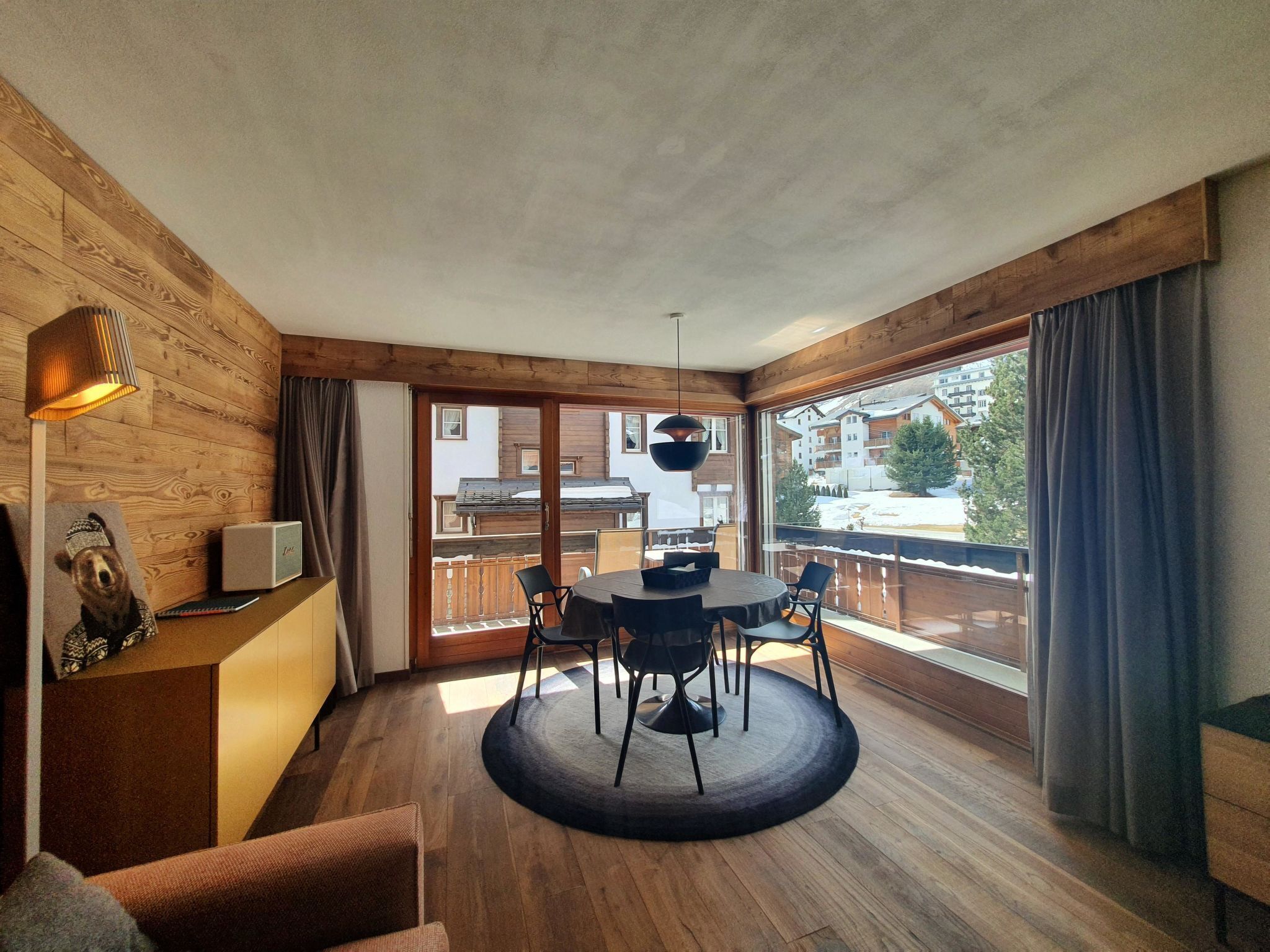 A modern interior with a wooden floor, a round table with chairs, a sofa, and a view of buildings through large windows.