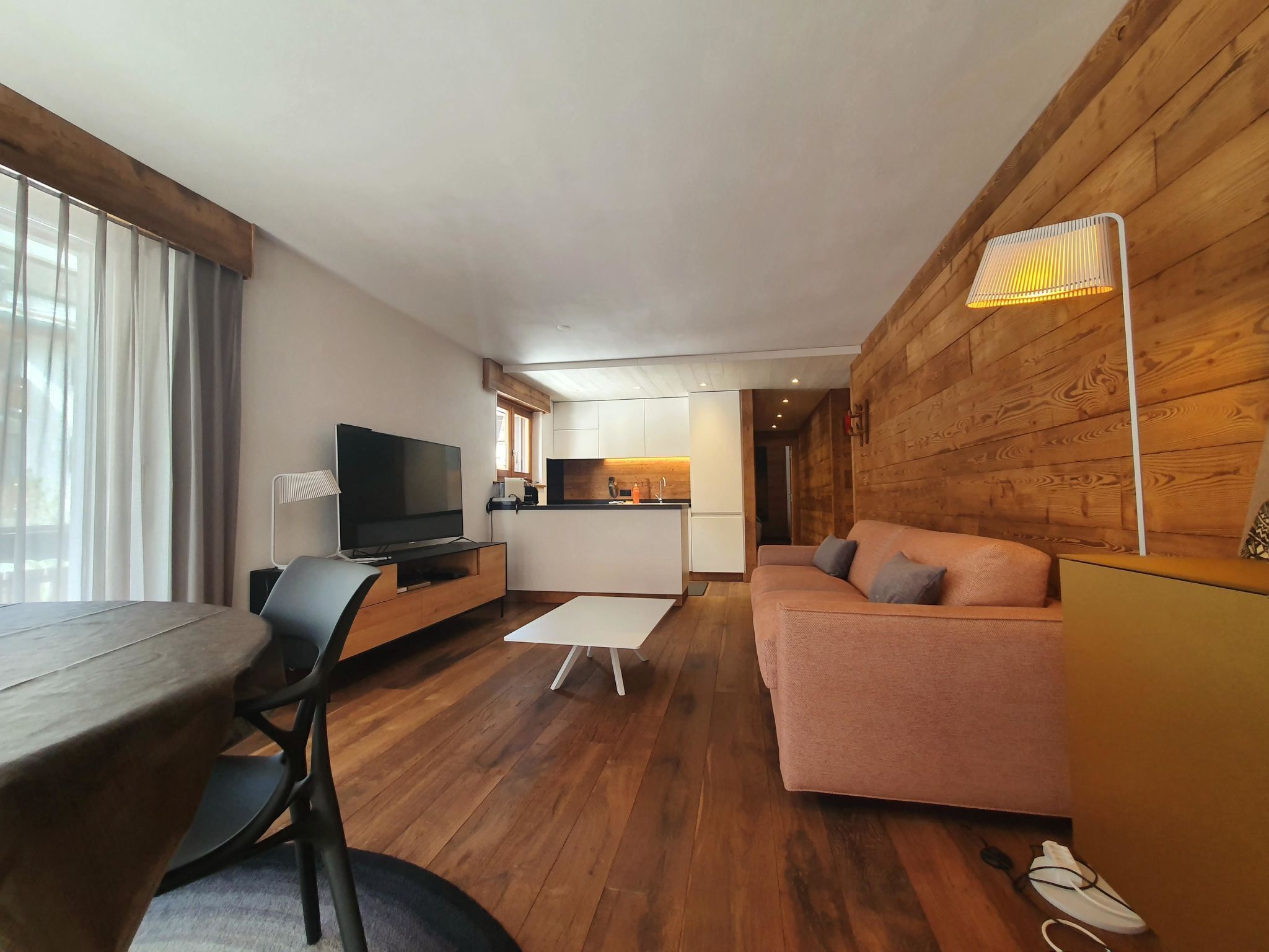 Una sala moderna con un televisor de pantalla plana, un sofá, una mesa de centro y un comedor con una cocina al fondo.