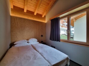 Une chambre avec un plafond en bois, un lit avec une literie blanche, une table de chevet, une fenêtre avec des rideaux et une vue sur un bâtiment à l’extérieur.