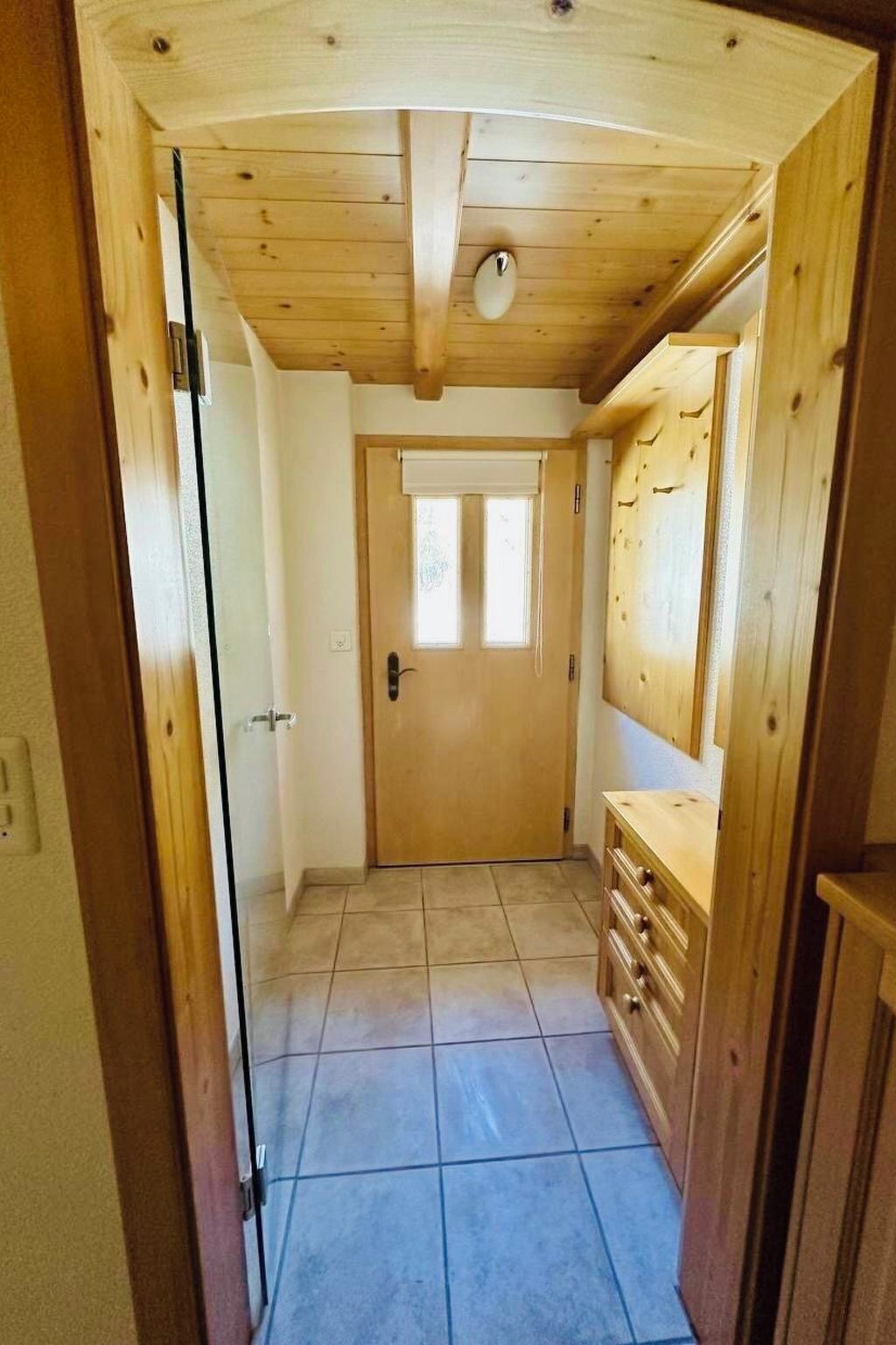 Un pasillo con techo de madera, una puerta con ventanal y piso de baldosas.