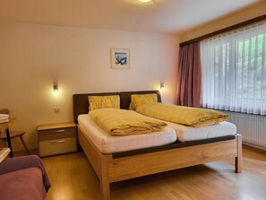 A bedroom with a wooden bed, bedside tables, a nightstand, a framed picture on the wall, and a window with curtains. - Apartments Atlas Appartement/Fewo Atlas 3 by Interhome (Saas-Grund)