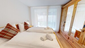Ett hotellrum med två sängar, vita sängkläder, kuddar med rutmönster, en garderob i trä och ett stort fönster med tunna, genomskinliga gardiner.