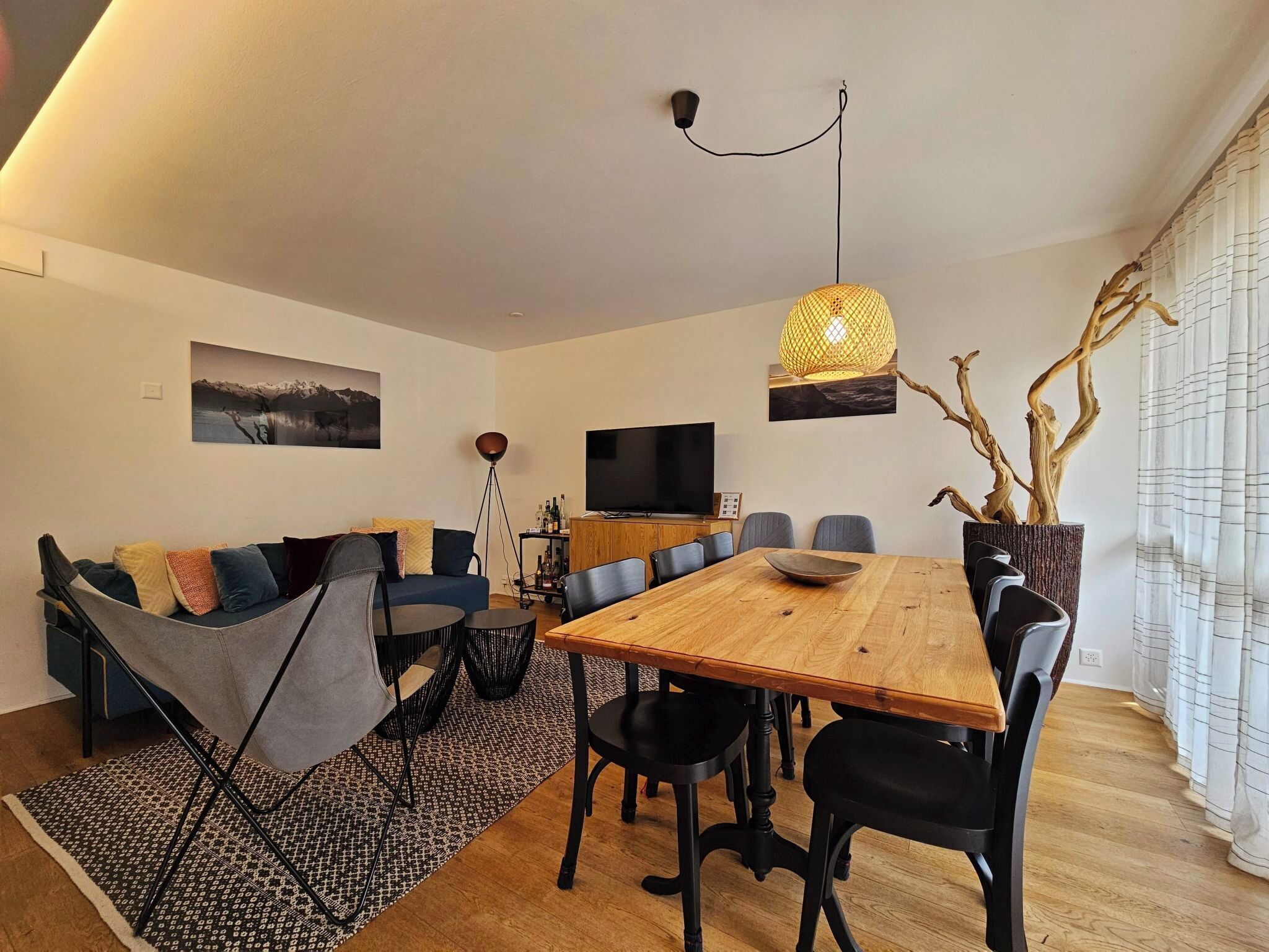 En moderne stue med et træspisebord, sorte stole, en sofa, et fjernsyn og et stort vindue med gardiner.
