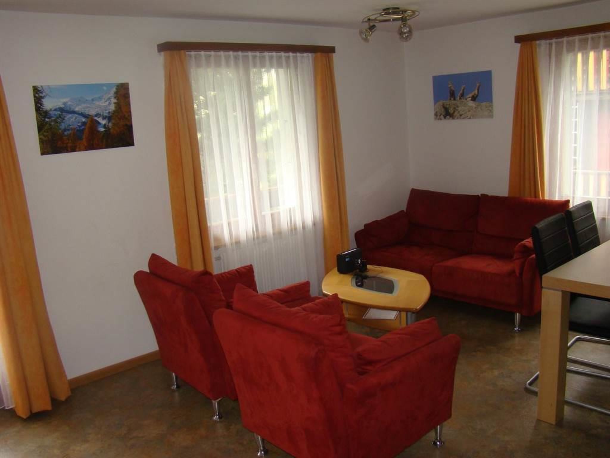 A room with a red sofa, a small table, and a wooden desk. There are two windows with curtains, and a painting on the wall.
