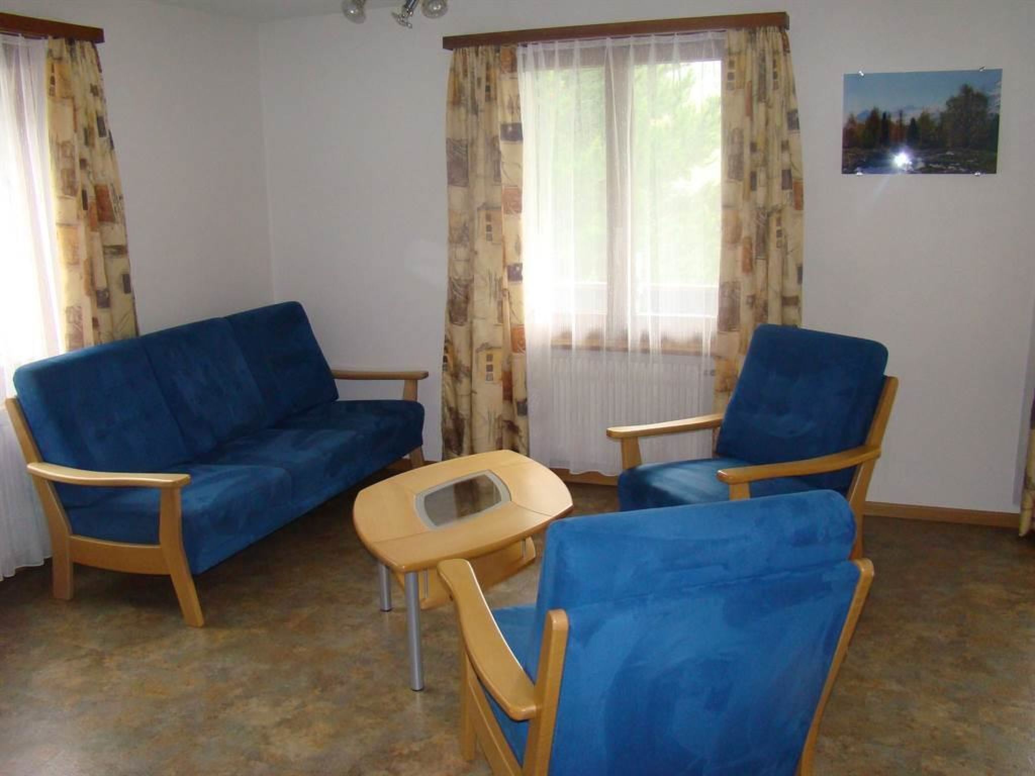 Una habitación con un sofá azul, un sillón azul, una mesita de madera y una ventana con cortinas.
