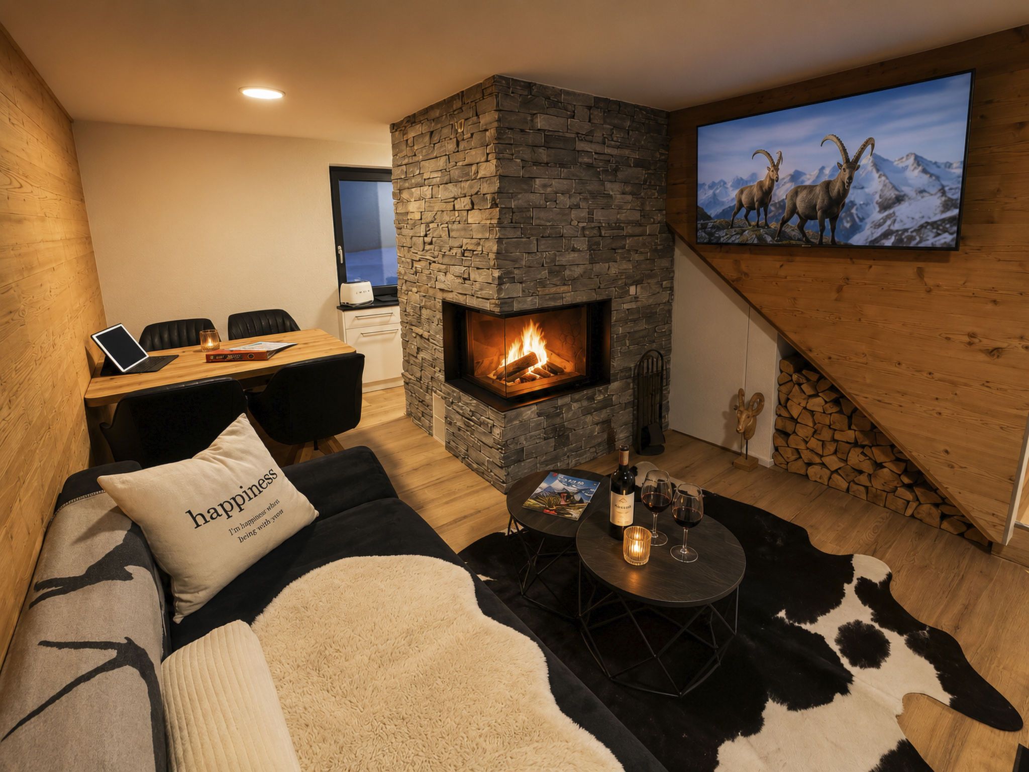 Un salon chaleureux avec une cheminée en pierre, un coin repas équipé d’un ordinateur portable et un téléviseur diffusant des images de chèvres de montagne.