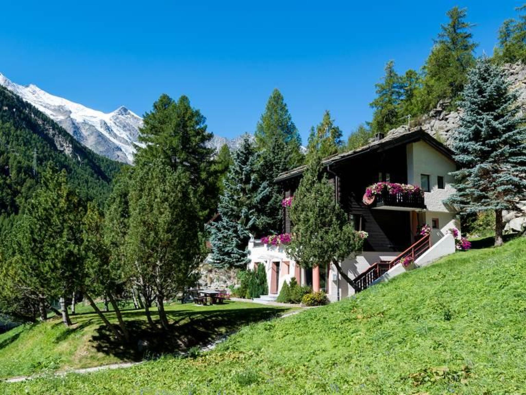 A mountain lodge with a balcony, surrounded by trees and snow-capped mountains.