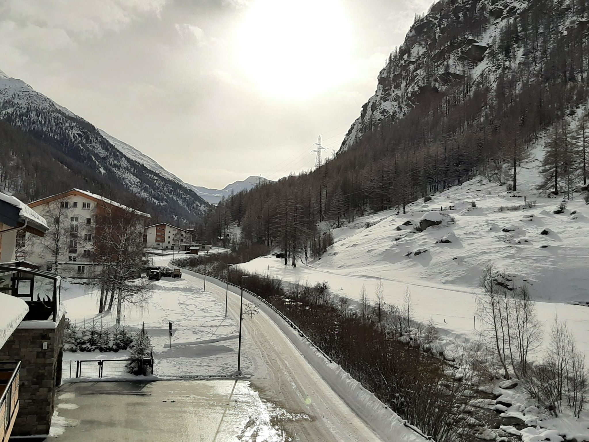 A snow-covered mountain road with buildings on the left, a clear sky, and a valley.