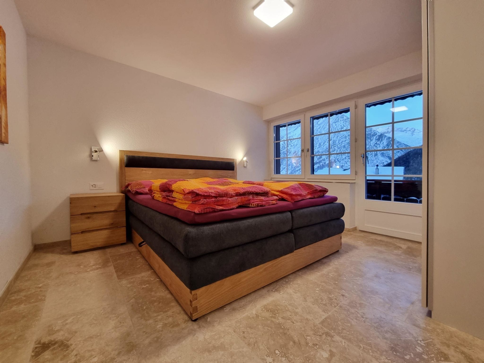 A bedroom with a bed, bedside tables, a window, and a view of snow-covered mountains.