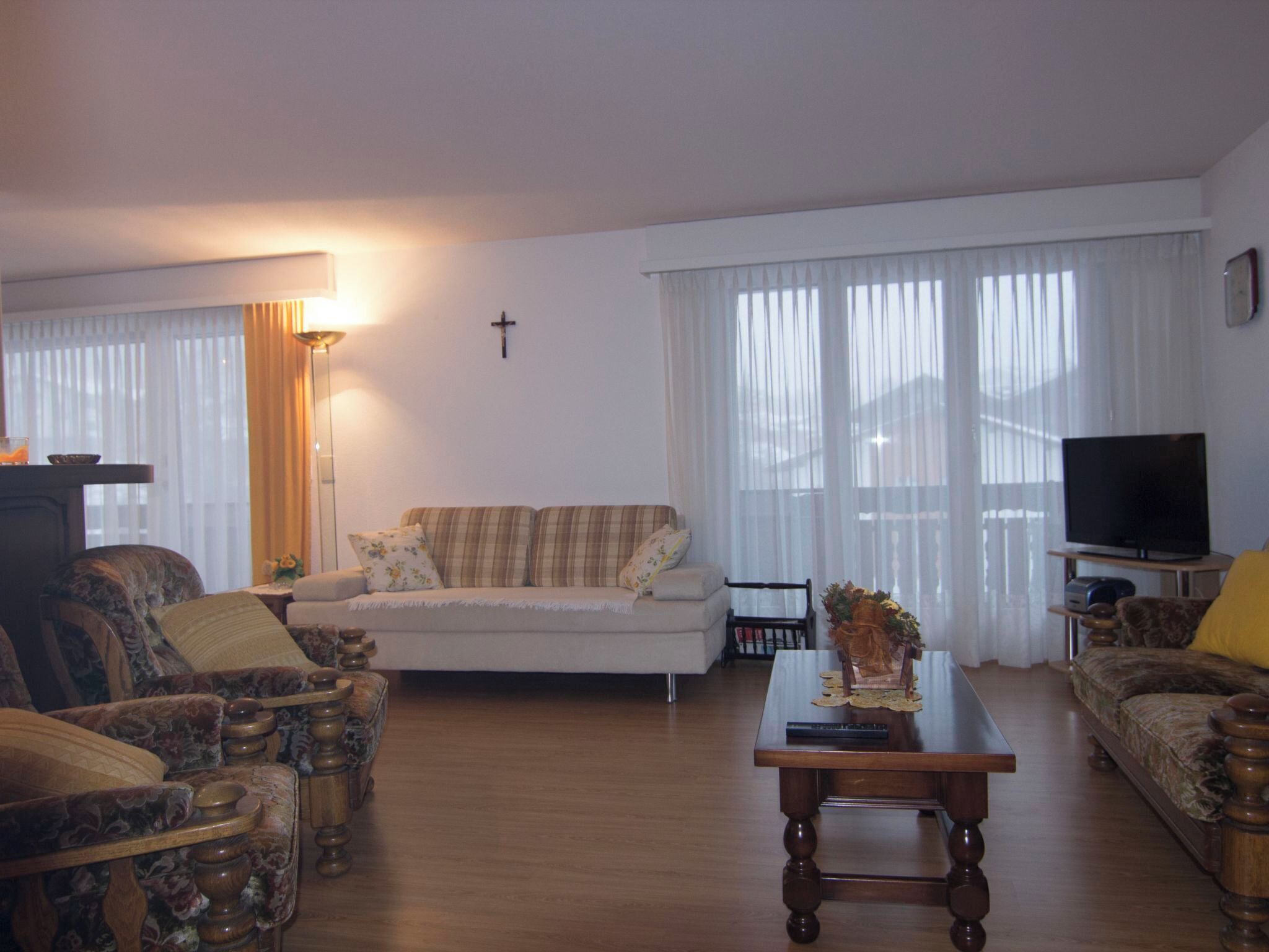 Een woonkamer met een bank, fauteuils, een salontafel, een televisie en een kruis aan de muur.