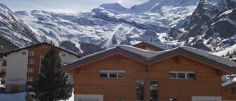 A wooden chalet with a mountain backdrop, featuring a clear sky and snow-covered peaks.