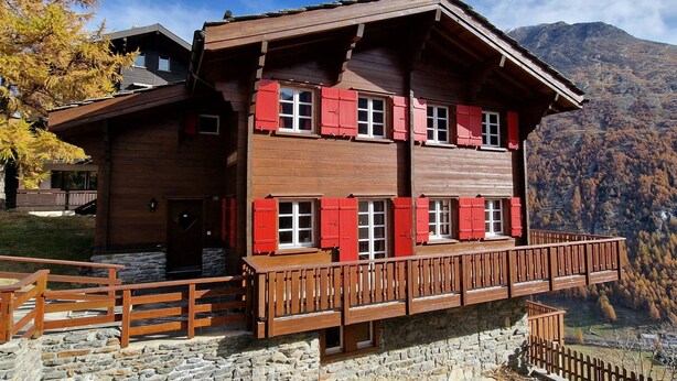 A multi-story wooden chalet with red shutters, stone foundation, and a balcony overlooking a mountainous landscape. - Les Mélèzes Appartement/Fewo 8-Bettwohnung by Interhome (Saas-Fee)