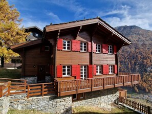 Un chalet en bois à plusieurs étages, avec des volets rouges, des fondations en pierre et un balcon donnant sur un paysage montagneux.