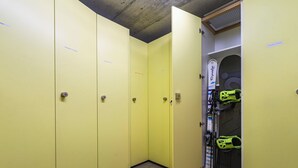 Um vestiário amarelo com vários armários, uma porta que dá para uma área de armazenamento com esquis, e um letreiro com a inscrição "Victoria's Home".