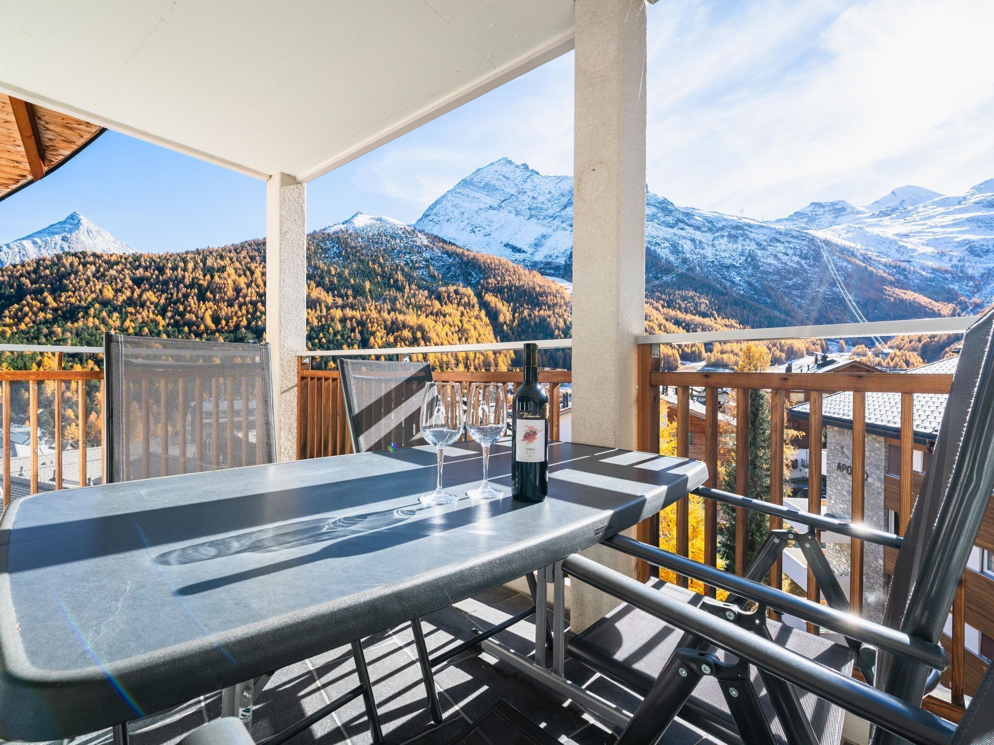 A balcony with a table and chairs, overlooking a mountainous landscape with autumn foliage.