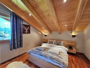 A bedroom with a wooden ceiling, a bed with a fluffy blanket, a window with curtains, and a wall-mounted light. - Aramis 5. OG Appartement/Fewo 6-Bettwohnung by Interhome (Saas-Fee)
