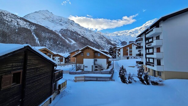 Un pueblo en la montaña cubierto de nieve con edificios de madera y modernos, una valla de madera y un cielo despejado.