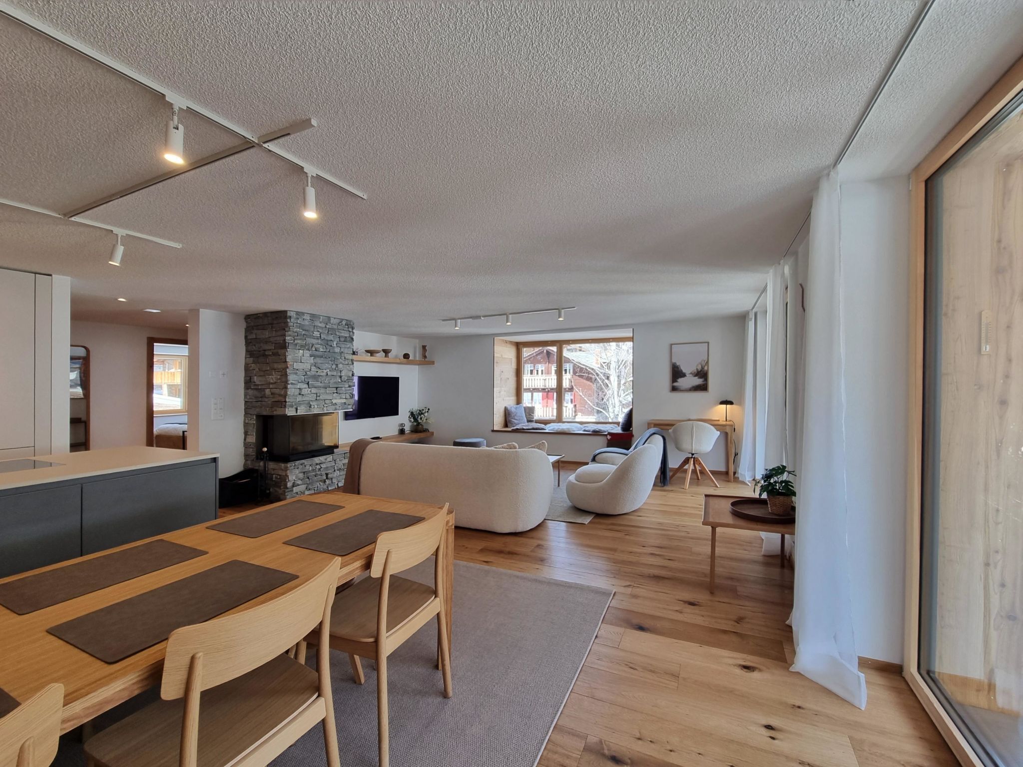 A modern living room with a stone fireplace, a dining area with a wooden table and chairs, and a large window with a view of buildings.