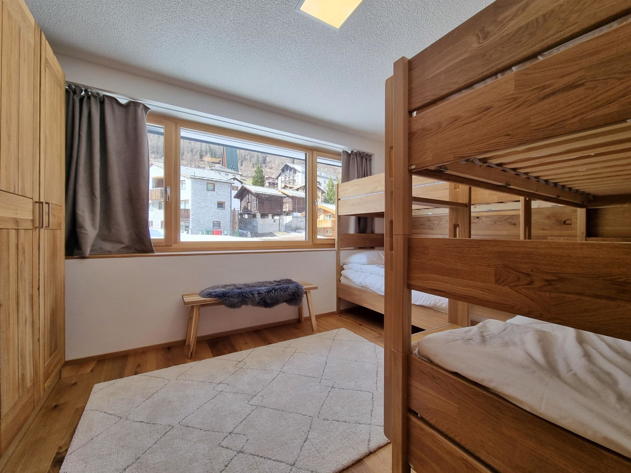 Une chambre avec un lit superposé, une fenêtre, une armoire en bois et une petite table recouverte d’un plaid en fausse fourrure.