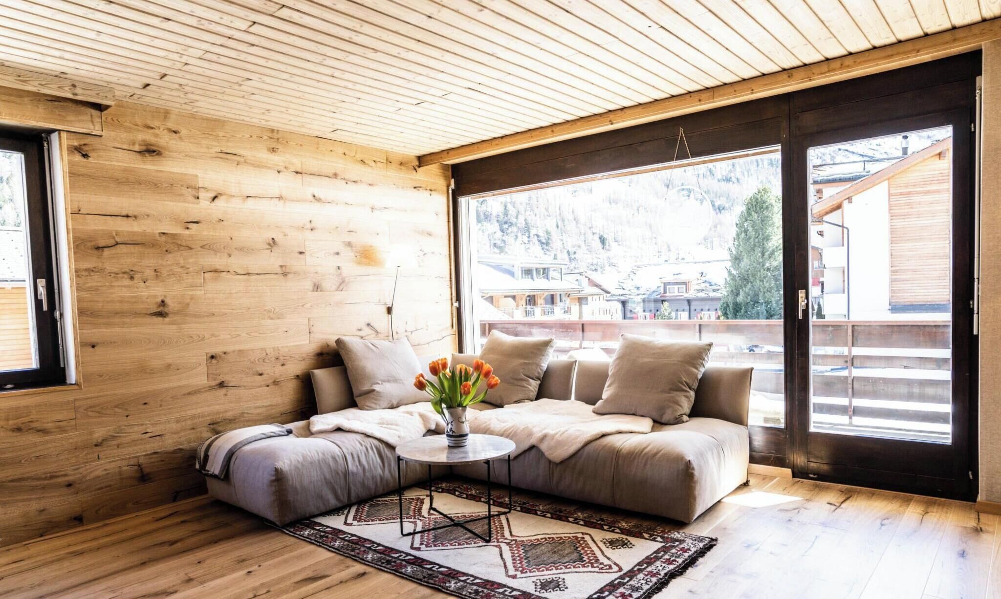 A cozy room with a wooden interior, a sofa, a small table, and a view of a snowy landscape through large windows.