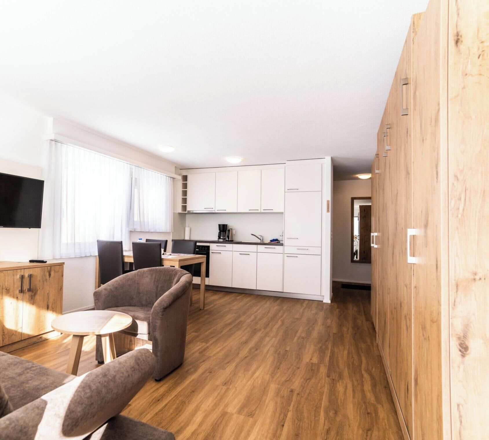 Una cocina moderna con armarios blancos, una zona de comedor con mesa y sillas, y suelo de madera.