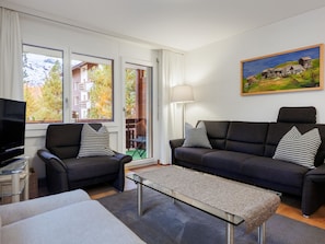 Ein modernes Wohnzimmer mit einer Couch, Sesseln, einem Glastisch und einem Fernseher.