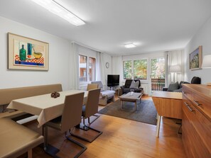 Ein modernes Wohnzimmer mit Essbereich, einem Flachbildfernseher und einem Balkon mit Blick auf Bäume.
