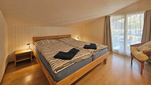 Une chambre avec un lit en bois, une table de chevet, une chaise et une vue sur les maisons par la fenêtre.
