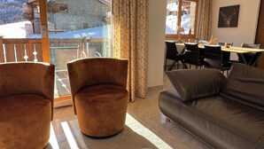 Ein gemütliches Wohnzimmer mit einer Couch, zwei Sesseln, einem Essbereich mit Tisch und Stühlen sowie Blick auf ein Bergchalet durch große Fenster.