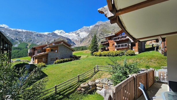 Blick auf Berge mit Holzzaun, Steinhaus und einem modernen Gebäude mit Balkonen.