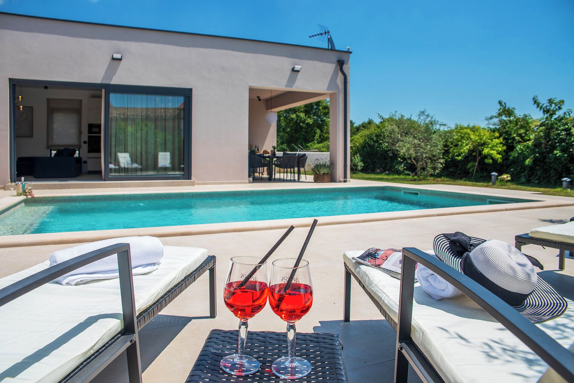 A modern poolside area with lounge chairs, a table with two glasses of red drink, and a view of a building with a pool.