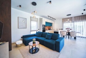 A modern living room with a sectional sofa, a dining area with a table and chairs, a kitchen in the background, and a large window with curtains. - Villa Lucia by Istrabook – Peace, Nature & Private Pool (Marcana)