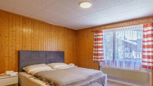 Makuuhuone, jossa on puupaneloitu seinä, harmaalla päätyosalla varustettu sänky, valkoiset vuodevaatteet ja ikkuna, jossa on punavalkoiset verhot.