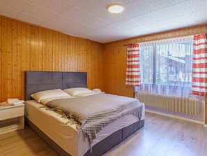 A bedroom with a wooden paneled wall, a bed with a grey headboard, white bedding, and a window with red and white curtains. - Haus Slalom Appartement/Fewo Apt #4 by Interhome (Saas-Fee)